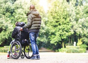 An adult male pushes a wheelchair holding another person outside surrounded by trees near a park