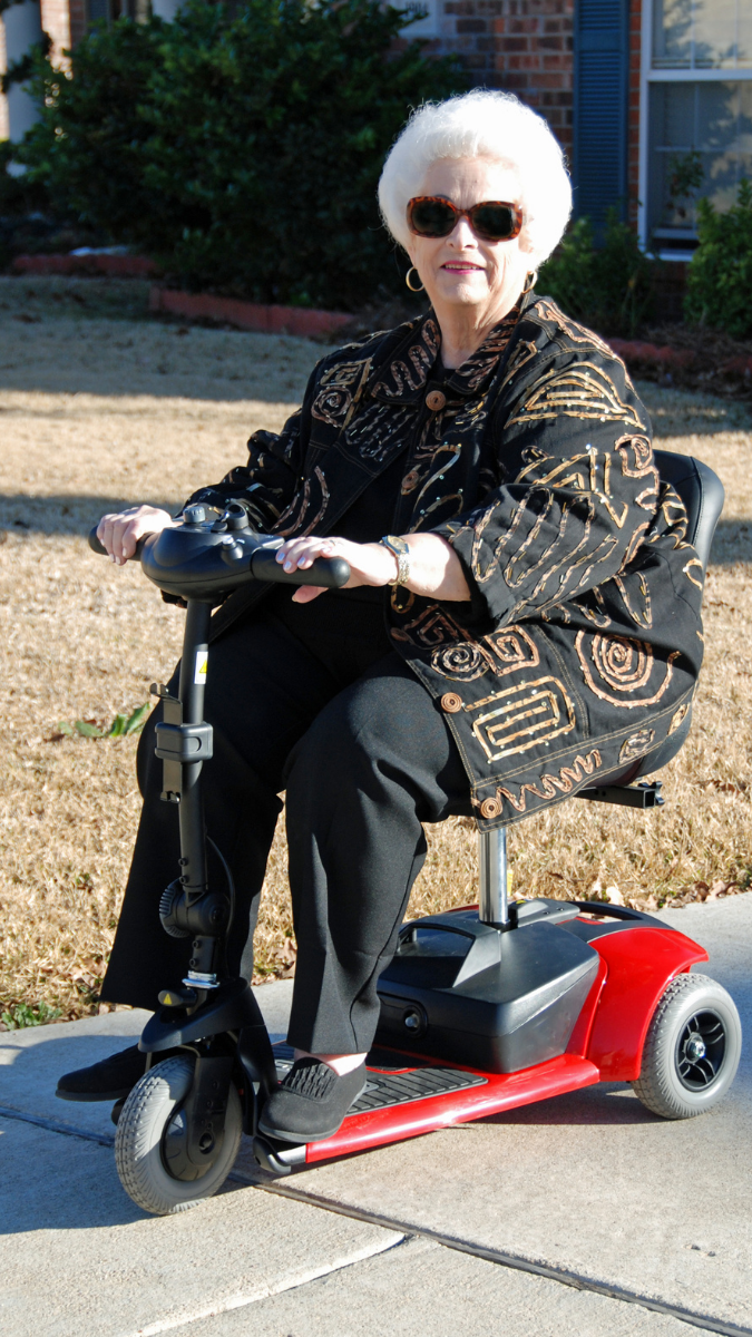 elderly woman wearing dark sunglasses on a mobility scooter while she rides down the sidewalk