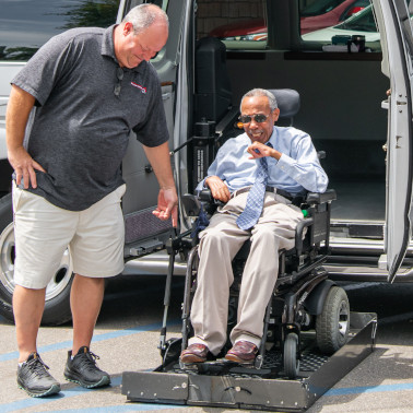elderly gentleman sits in a powerchair on top of a platform lift system attached to a van. A man stands next to him, leaning over to assist.